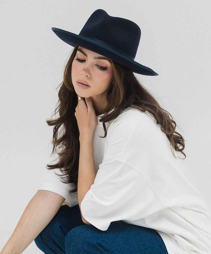 Woman wearing a navy fedora hat with a band, a white t-shirt and blue pants, sitting against a plain background #color_navy