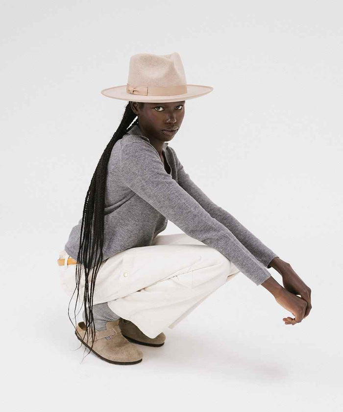 Woman wearing an oatmeal colored fedora hat with a band, a grey shirt, white pants and brown slides, sitting against a plain background #color_oatmeal