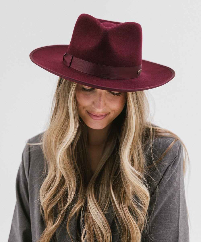 Woman wearing a wine colored fedora hat with a band and a grey blazer, looking down against a plain background #color_wine