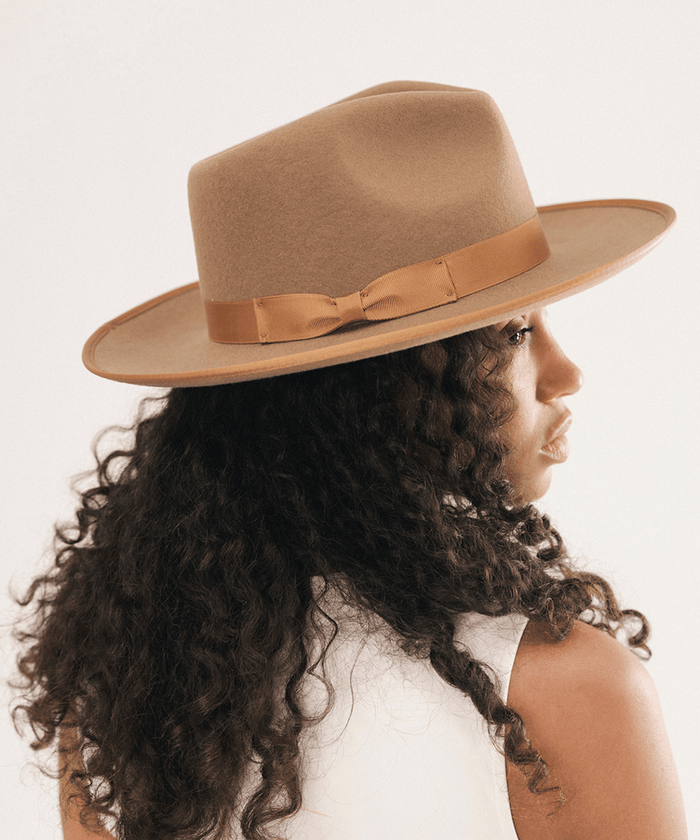 A woman with long, dark curly hair is shown from the back and side profile, wearing a wide-brim fedora hat in a warm camel brown with a matching brown ribbon band and bow. The background is a clean, off-white studio setting #color_brown