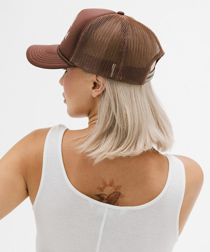Woman wearing a brown trucker hat and a white top, facing sideways against a plain background  #color_chocolate brown