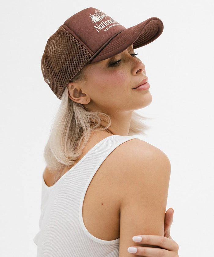Woman wearing a brown trucker hat with 'National Park' logo and a white top, posing sideways against a plain background  #color_chocolate brown