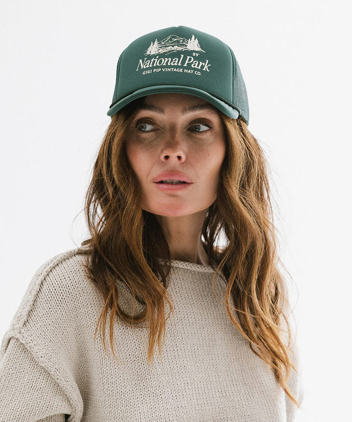 Woman wearing a forest green colored trucker hat with 'National Park' logo and a grey sweatshirt against a plain background  #color_forest green
