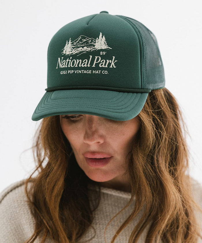 Woman wearing a forest green colored trucker hat with 'National Park' logo against a plain background  #color_forest green