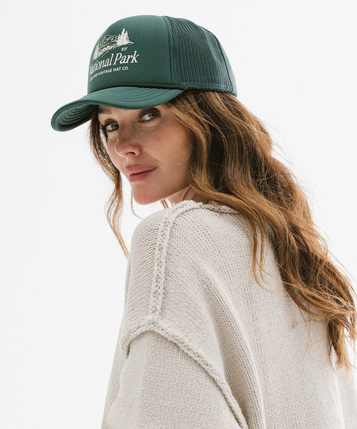 Woman wearing a forest green colored trucker hat with 'National Park' logo and a grey sweatshirt, posing sideways against a plain background  #color_forest green
