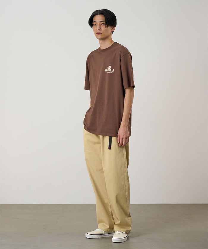 Model wearing a Natural Dyed Gramicci Pant Straight Fit in chino and brown shirt on a light-tone background.