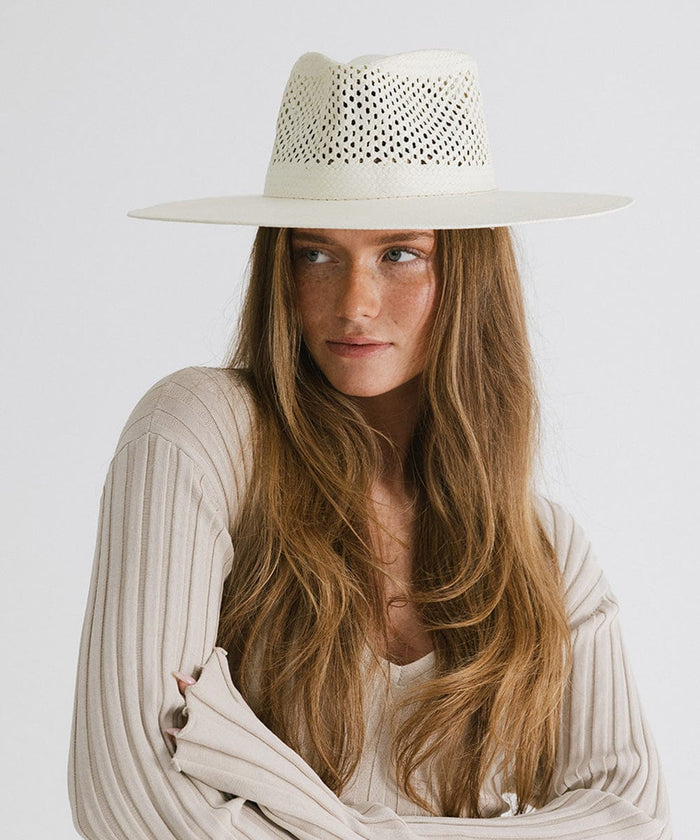 Woman wearing a natural white fedora hat with a wide brim and intricate venting details, and a cream top, looking sideways against a plain background #color_natural white