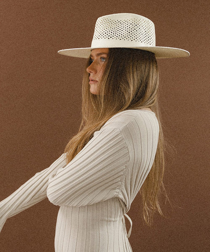 Woman wearing a natural white fedora hat with a wide brim and intricate venting details, and a cream top, facing sideways against a brown background #color_natural white
