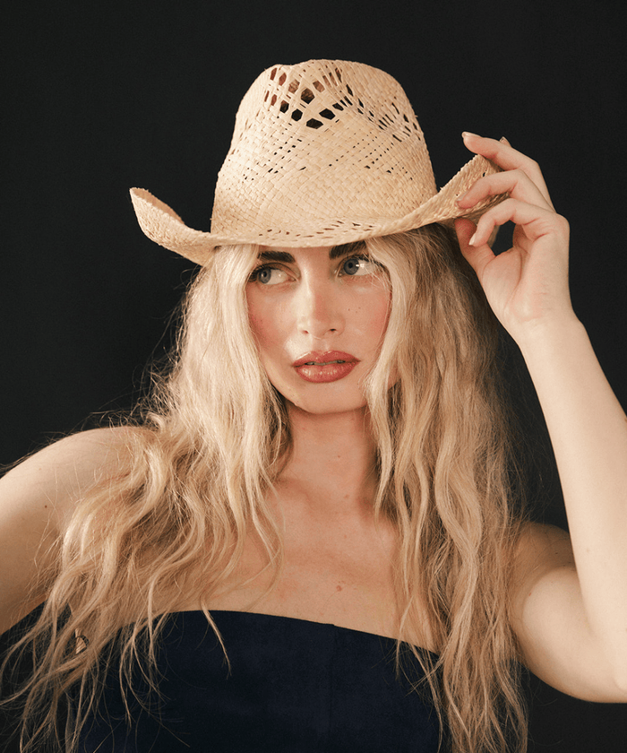 Woman wearing a natural colored straw fedora hat and dark clothing against a plain background #color_natural