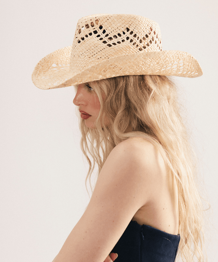 Woman wearing a natural colored straw fedora hat and dark clothing against a plain background #color_natural