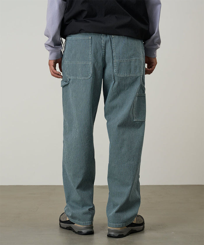 Back view of a model wearing navy Gramicci Overdyed Hickory Work Pants and a black vest on a light-tone background.