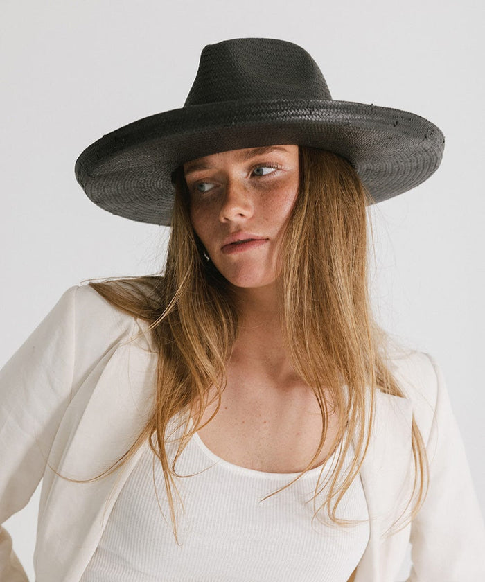 Woman wearing a black pencil brim straw hat and a white top with a white blazer against a plain background #color_black