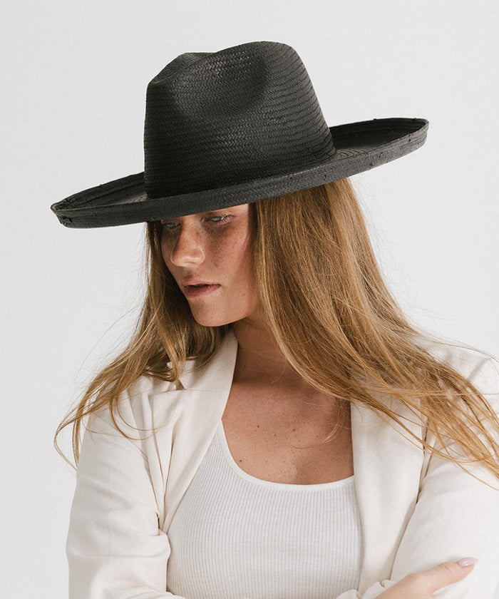 Woman wearing a black pencil brim straw hat and a white top with a white blazer, looking sideways against a plain background #color_black