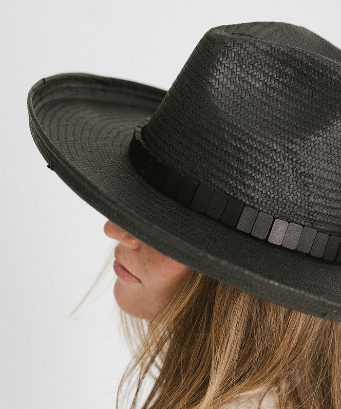 Close up image of a woman wearing a black pencil brim straw hat with a hat band against a plain background #color_black