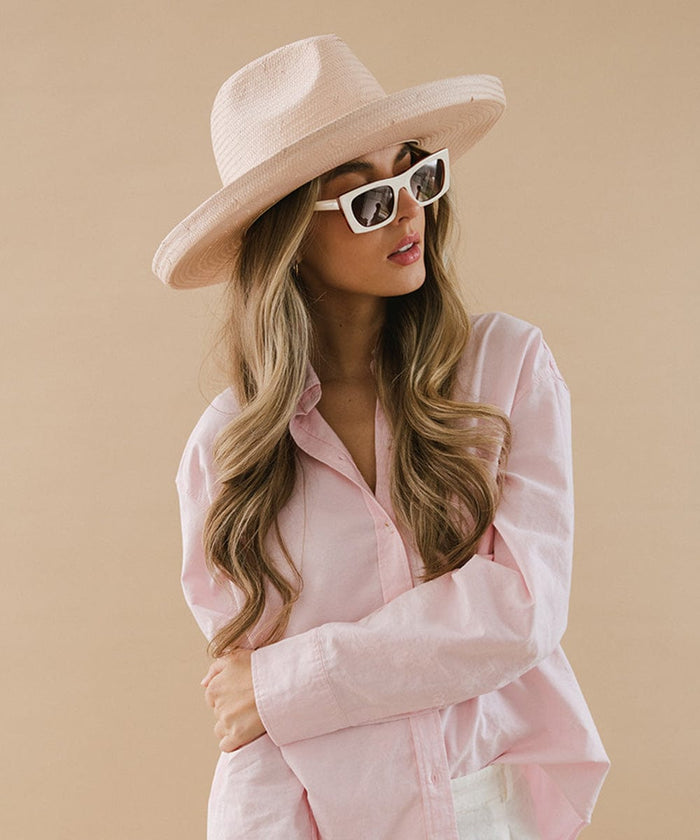Woman wearing a light pink pencil brim straw fedora hat, sunglasses, and a pink shirt, looking sideways against a cream background #color_limited edition light pink