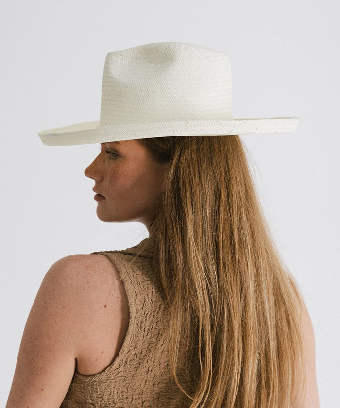 Woman with long hair wearing a white pencil brim fedora hat and a brown top, facing sideways against a plain background  #color_white