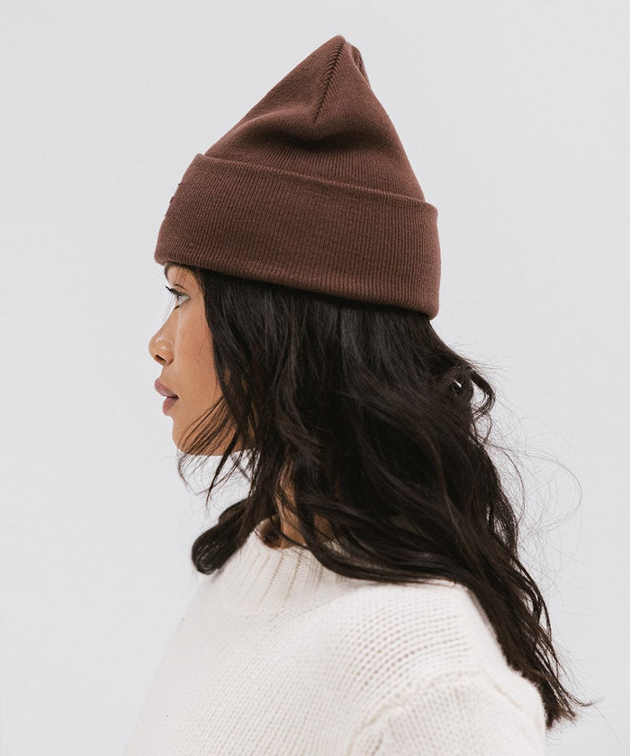Woman wearing a chocolate colored knit beanie and a white sweater, facing sideways against a plain background #color_chocolate