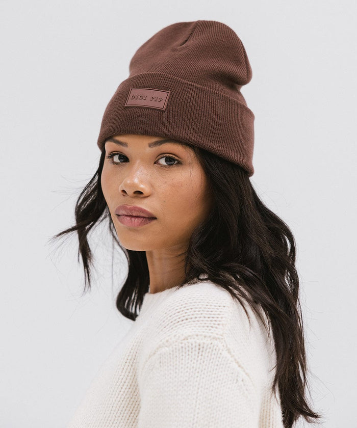 Woman wearing a chocolate colored knit beanie and a white sweater, posing sideways against a plain background #color_chocolate