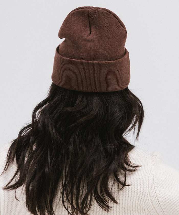 Woman wearing a chocolate colored knit beanie and a white sweater, facing behind against a plain background #color_chocolate