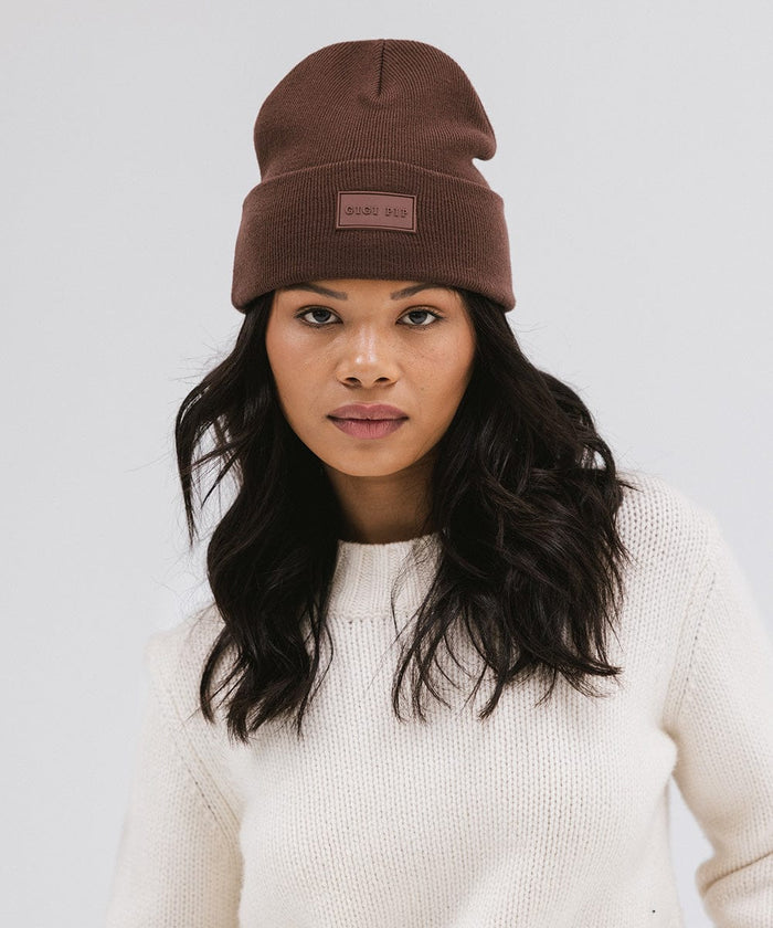 Woman wearing a chocolate colored knit beanie and a white sweater against a plain background #color_chocolate