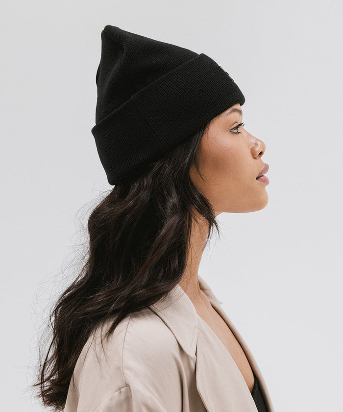 Woman wearing a black knit beanie and a beige coat, facing sideways against a plain background #color_black