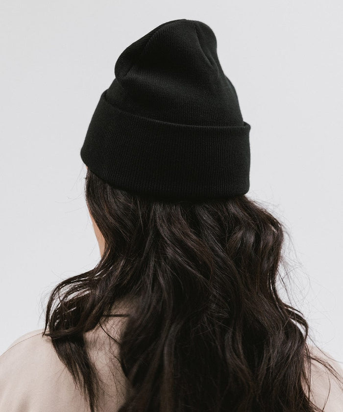 Woman wearing a black knit beanie and a beige coat, facing behind against a plain background #color_black