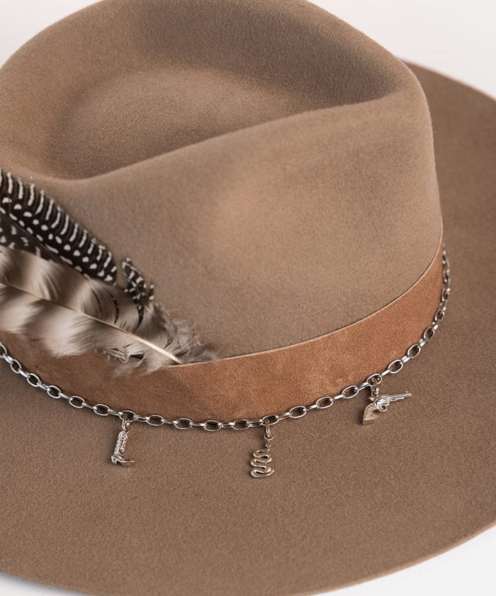 A brown hat with a band, two hat feathers and a silver chain with gold boot hat charm, snake hat charm and pistol hat charm on a plain background #color_gold