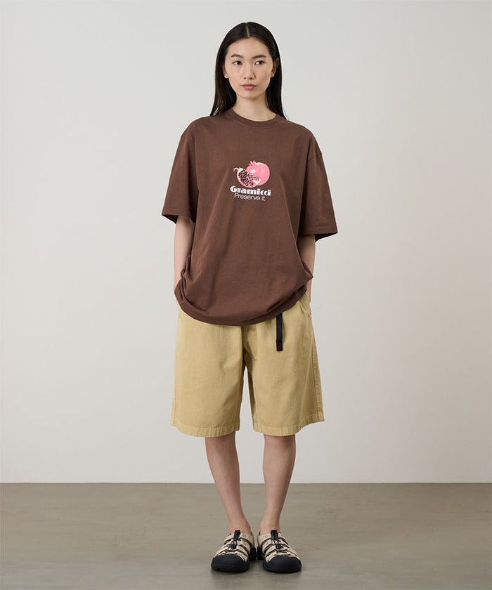 Model wearing a brown Gramicci Pomegranate Tee with a a graphic and beige shorts on a light tone background.