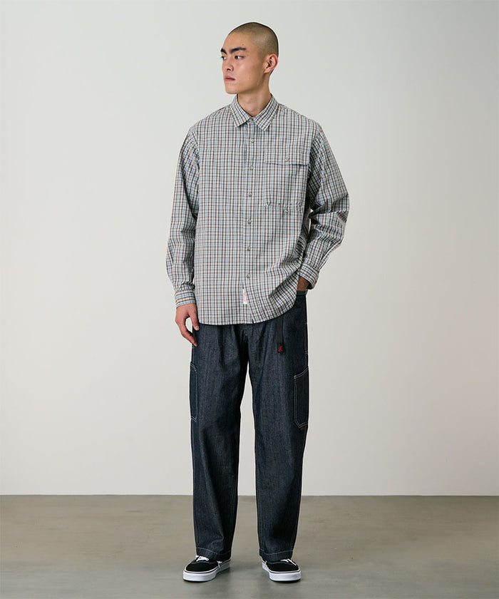 Model wearing a brown plaid Gramicci Poplin Millwood Shirt and blue pants on a light-tone background.
