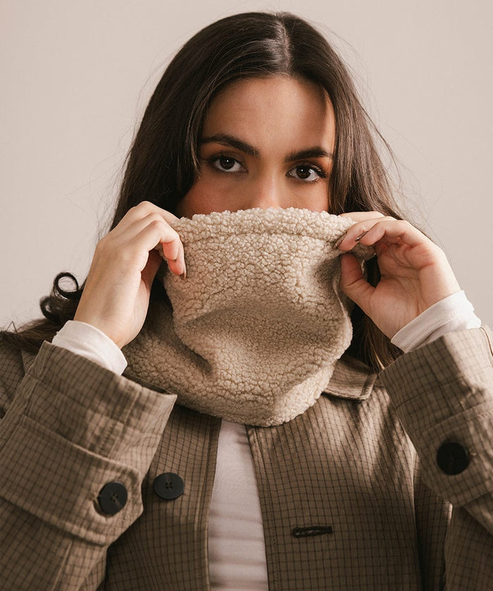 Woman wearing a brown coat with a textured scarf, holding it up to her face. #color_cream