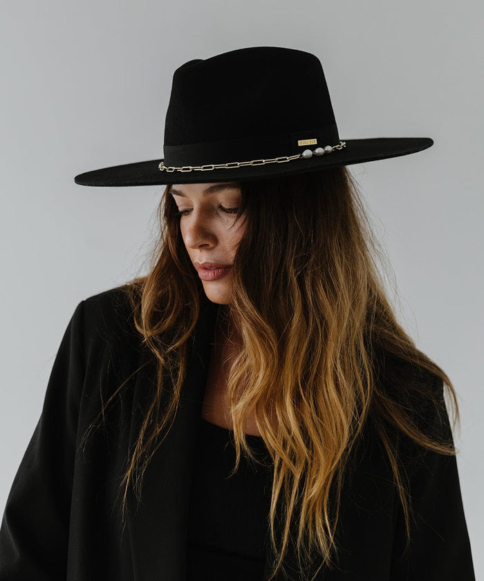 Woman wearing a black wide brim fedora hat with a gold chain detail around the base of the crown and a black coat, looking sideways against a plain background  #color_black