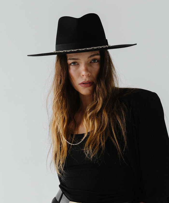 Woman wearing a black wide brim fedora hat with a gold chain detail around the base of the crown and a black top with a black coat against a plain background  #color_black