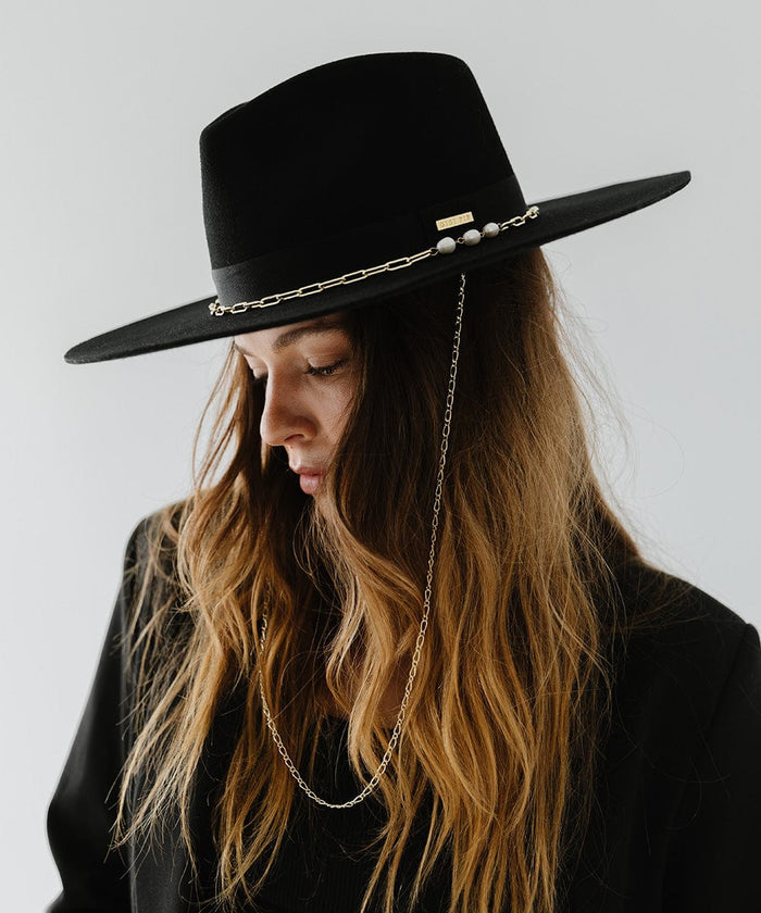 Woman wearing a black wide brim fedora hat with a gold chain detail around the base of the crown and a black coat, looking down against a plain background  #color_black