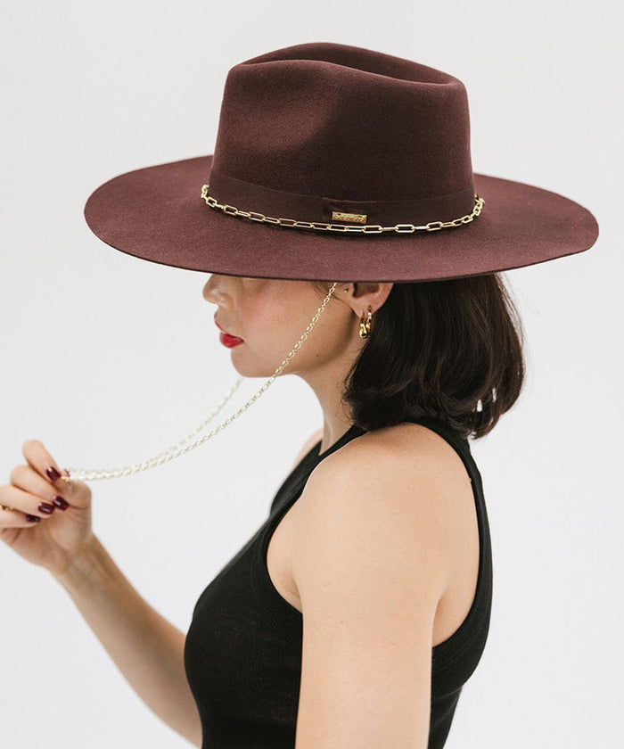 Woman wearing a dark cherry colored wide brim fedora hat with a gold chain detail around the base of the crown and a black top, facing sideways against a plain background #color_dark cherry