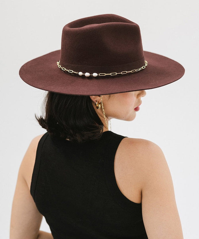 Woman with short hair wearing a dark cherry colored wide brim fedora hat with a gold chain detail around the base of the crown and a black top, looking sideways against a plain background #color_dark cherry