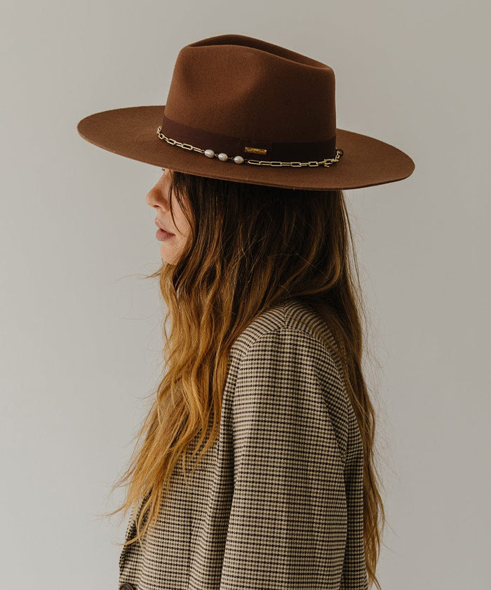 Woman wearing a dark oak colored wide brim fedora hat with a gold chain detail around the base of the crown and a coat, facing sideways  against a plain background  #color_dark oak
