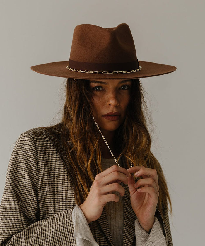Woman wearing a dark oak colored wide brim fedora hat with a gold chain detail around the base of the crown and a coat, posing against a plain background  #color_dark oak