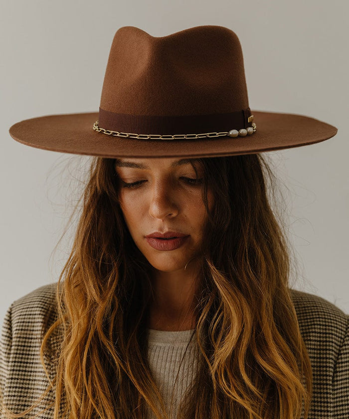 Woman wearing a dark oak colored wide brim fedora hat with a gold chain detail around the base of the crown against a plain background  #color_dark oak