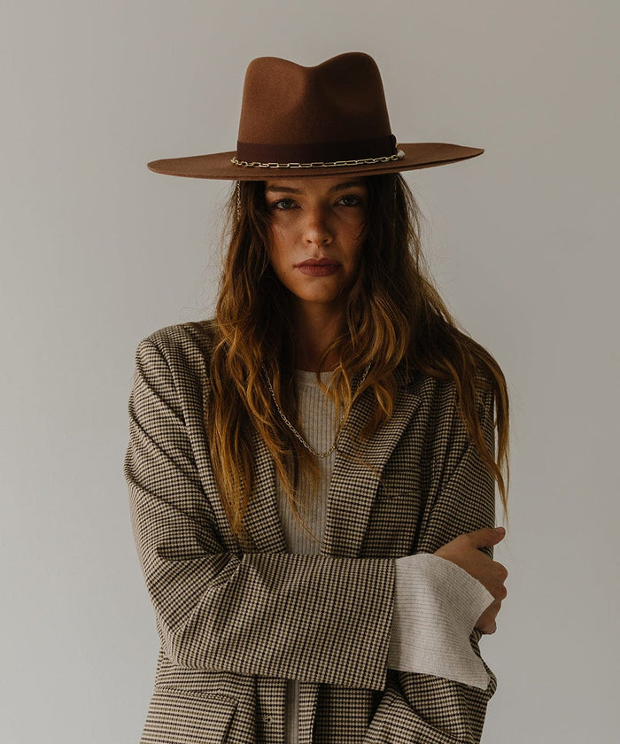 Woman wearing a dark oak colored wide brim fedora hat with a gold chain detail around the base of the crown and a coat against a plain background  #color_dark oak