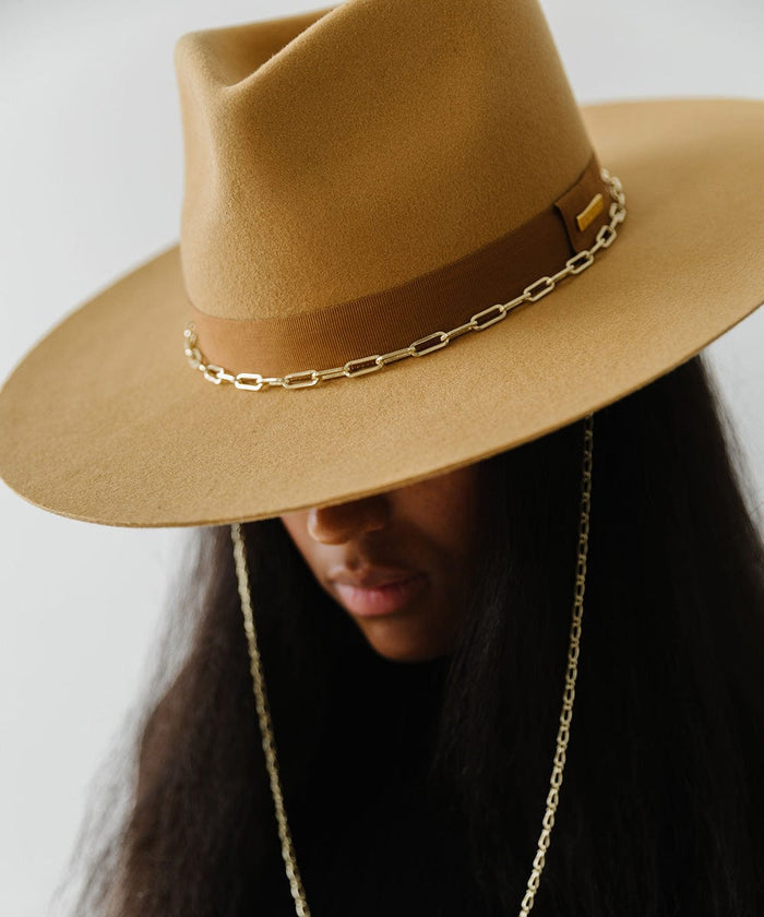Person wearing a honeycomb  colored wide brim fedora hat with a band and a gold chain detail around the base of the crown against a plain background #color_honeycomb