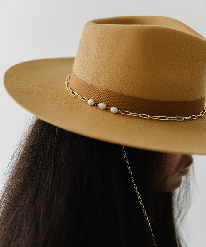 Woman wearing a honeycomb colored wide brim fedora hat with a band and a gold chain detail around the base of the crown, facing sideways against a plain background #color_honeycomb