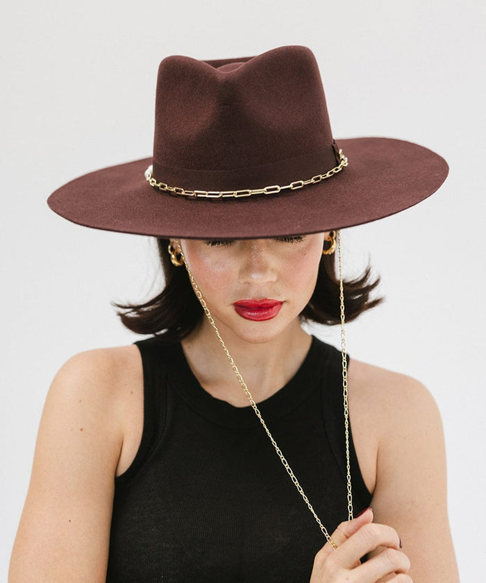Woman wearing a dark cherry colored wide brim fedora hat with a gold chain detail around the base of the crown and a black top, looking down against a plain background #color_dark cherry