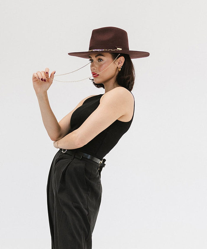 Woman wearing a dark cherry colored wide brim fedora hat with a gold chain detail around the base of the crown, a black top and black pants against a plain background #color_dark cherry