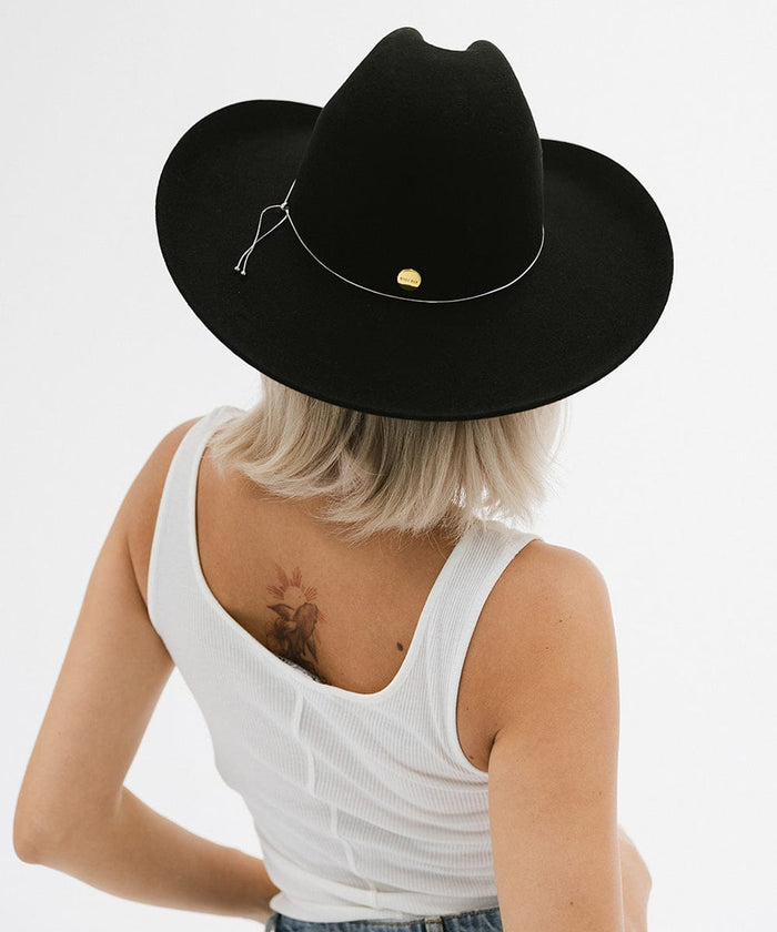 Woman with short hair wearing a black wide brim hat with a hat band and a white top against a plain background #color_black