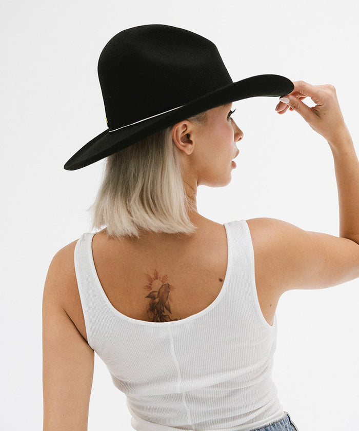 Woman with short hair wearing a black wide brim hat with a hat band and a white top, posing sideways against a plain background #color_black