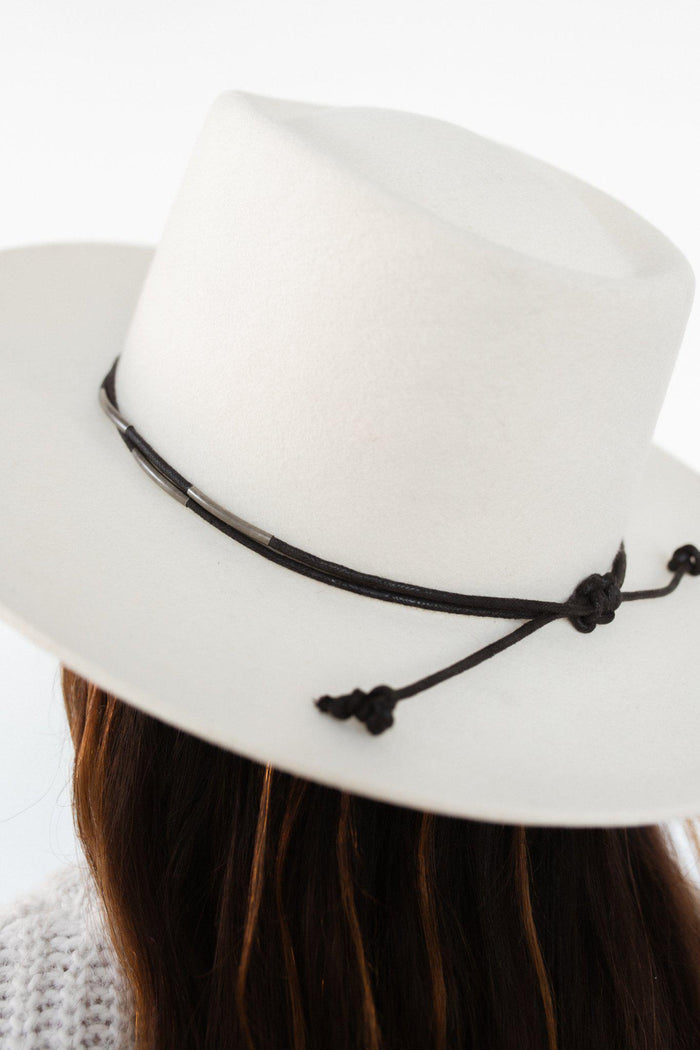 Close up image of a woman wearing a white hat with a black rope band with beads facing behind against a plain background #color_black