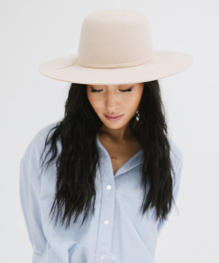 Woman wearing a cream open crown hat with a hat band and a blue shirt against a plain background  #color_cream