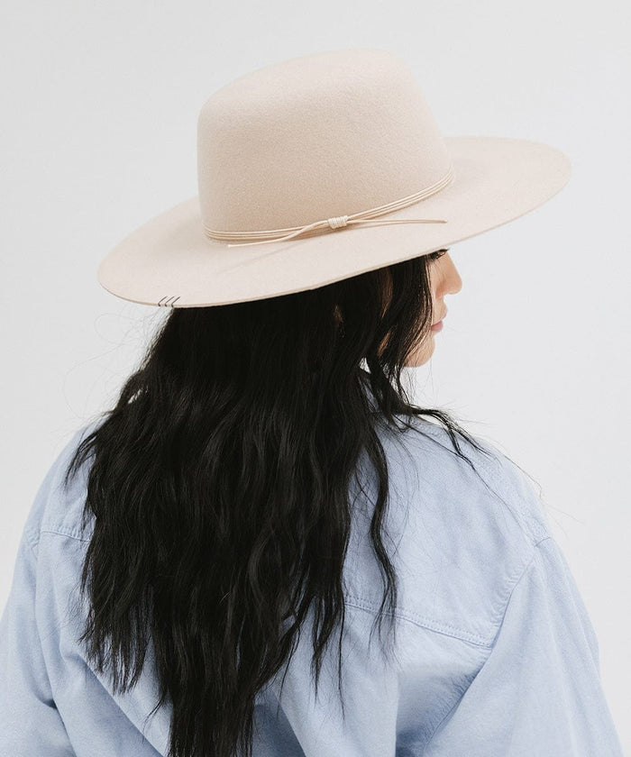 Woman with long black hair wearing a cream open crown hat with a hat band and a blue shirt, looking sideways against a plain background  #color_cream