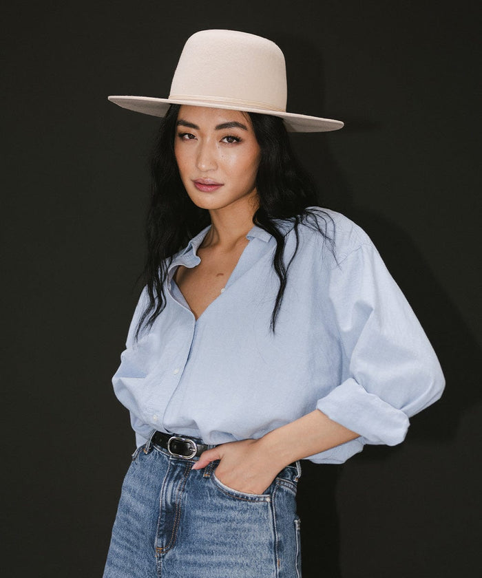 Woman wearing a cream open crown hat with a hat band, a blue shirt, black belt, and blue jeans against a dark grey background  #color_cream