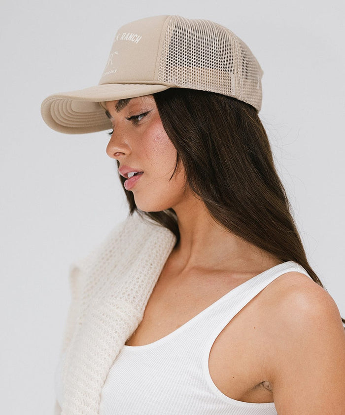 Woman wearing a tan cap with 'Saddleback Ranch' text and logo, and a white top, posing sideways against a plain background  #color_tan